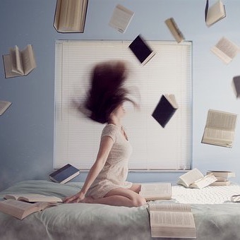 junge Frau kniet auf dem Bett mit fliegenden Haaren und Büchern die um sie herumwirbeln